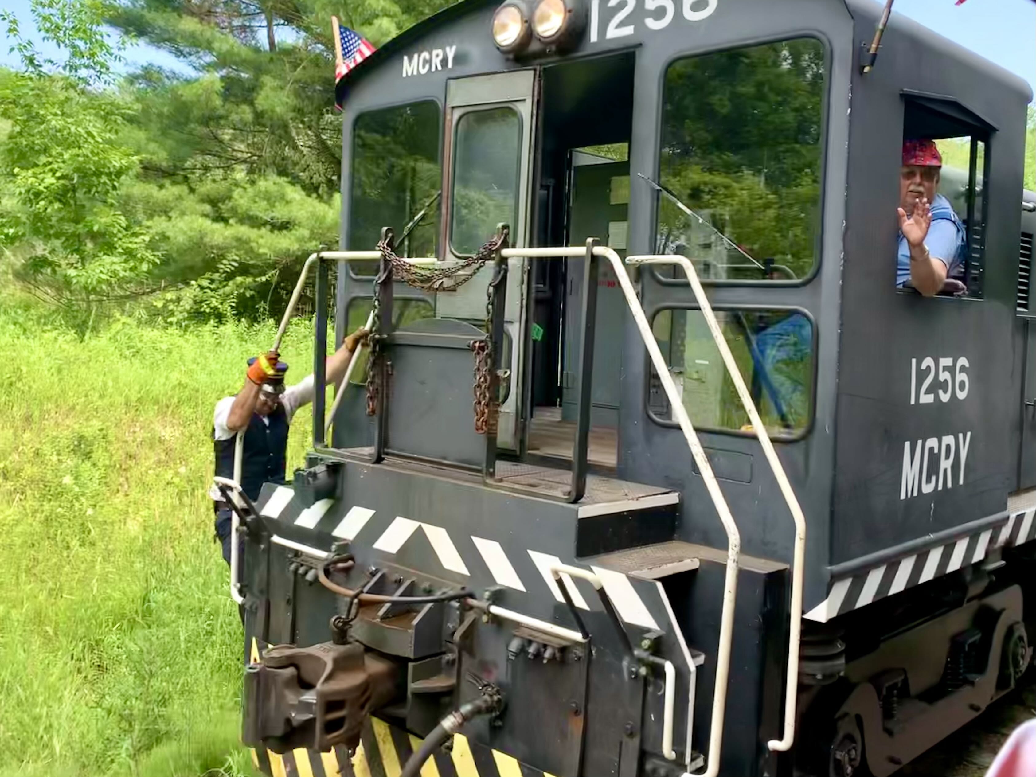 This image shows two people on a black train engine labeled "MCRY 1256." One person, wearing gloves and a safety vest, is climbing up the side of the train. Another person, sitting in the cab, waves out of the window. The train is moving through a green, forested area, and an American flag is displayed on the top corner of the train.