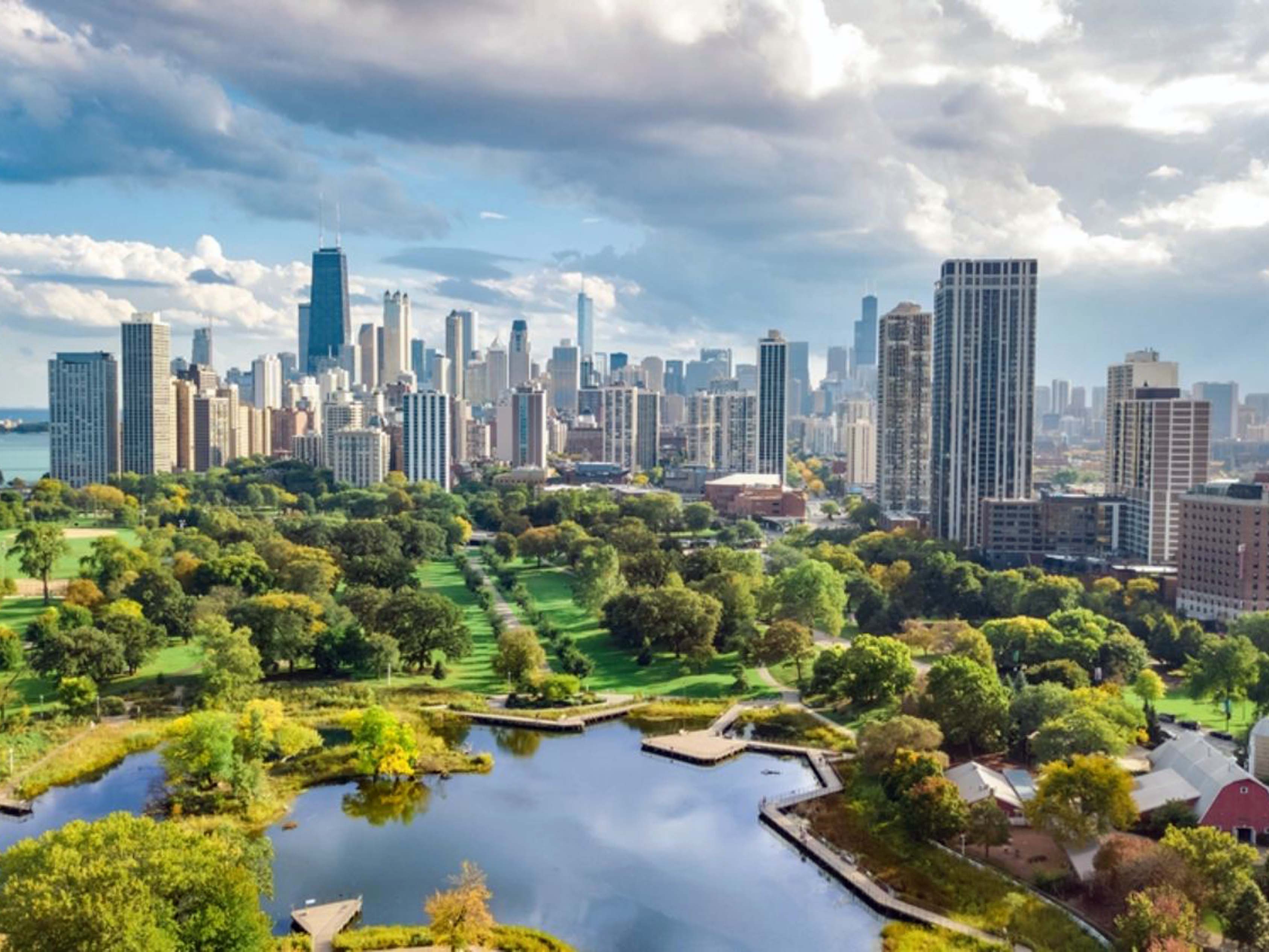 The image shows a wide-angle aerial view of downtown Chicago with its iconic skyline, including prominent skyscrapers like the Willis Tower and John Hancock Center. In the foreground is a lush, green park area featuring trees, ponds, and walking paths, which appears to be part of Lincoln Park. To the left, Lake Michigan is visible under a partly cloudy sky. The blend of urban architecture and natural greenery creates a striking contrast in this scenic shot of the city.